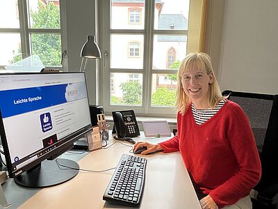 Anne Badmann an ihrem Schreibtisch im Büro. Sie trägt einen roten Pullover, lächelt und hat die rechte Hand auf er Computer-Mouse liegen. Vor ihr steht auf dem Schreibtisch ein großer Monitor, auf dem wiederum die Webseite Leichte-Sprache.online geöffnet ist. Davor liegt eine Computer-Tatstatur. Im Hintergrund ist ein Fenster.