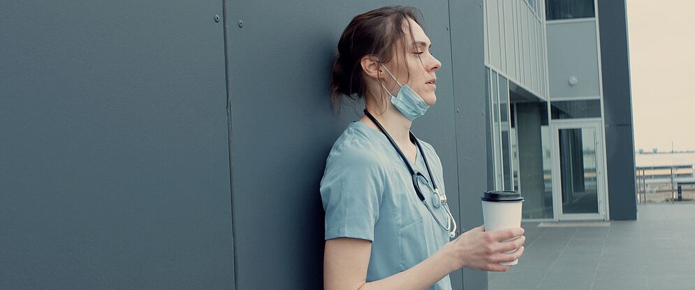 Eine Frau mit einem Stethoskop um den Hals und in Arzt- oder Pflegerkleidung lehnt an einer Hauswand mit einem Kaffeebecher in der Hand, der Maske unterm Kinn und einem müden Gesichtsausdruck. 